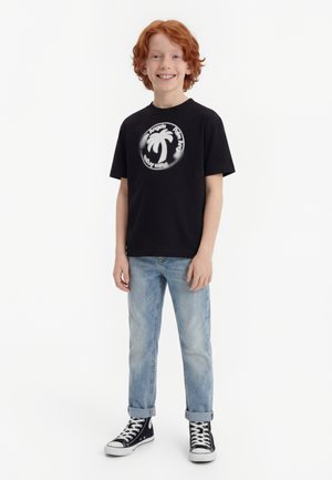 Χαμογελαστό αγόρι με κόκκινα μαλλιά που στέκεται, φορώντας μαύρο t-shirt με σχέδιο φοίνικα, ανοιχτόχρωμα τζιν με γυρισμένα μπατζάκια και μαύρα παπούτσια υψηλού τύπου.