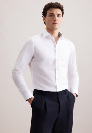 Mann mit lockigem Haar, der ein weißes Hemd mit Knöpfen und dunkle Hose trägt, steht mit den Händen in den Taschen vor einem schlichten Hintergrund.
