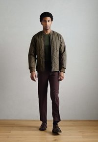 Jaqueta bomber almofadada verde-oliva por cima de uma camisa verde escura, combinada com calças castanhas e sapatos de couro escuro, num cenário minimalista.