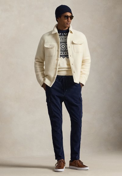 Muž nosiaci tmavomodrú čiapku, slnečné okuliare, krémový sveter so vzorom a bundu, tmavomodré nohavice typu cargo a hnedé tenisky s bielymi podrážkami.