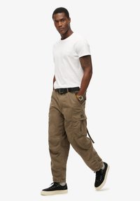 Cargo-broek in kaki, gemaakt van stevig materiaal, voorzien van zijzakken. Gepaard met een strakke witte t-shirt en zwarte sneakers.