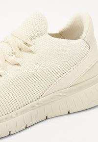 Cream textured sneaker featuring a ribbed knit upper, flat laces, and a chunky cushioned sole with subtle branding on the side.