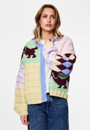 Kobieta ubrana w wielokolorowy, dziergany kardigan z wzorami psów i diamentów, na białym topie i niebieskich dżinsach, stojąca na białym tle.