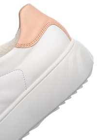 Bílá pata tenisky s tlustou podrážkou a světle broskvovým koženým akcentem na zadním límci, zobrazující detaily vyšívání.