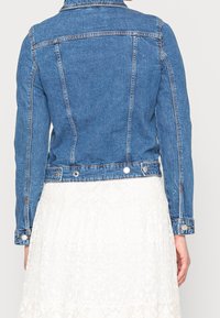 Jeansjacka i mellanblå tvätt med en figurnära design, knappade ärmslut och subtila sömnadsdetaljer, kombinerad med en vit spetskjol.