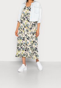 Robe longue fleurie en crème avec des fleurs bleues, jaunes et noires, assortie à une légère veste en denim et des baskets compensées blanches.