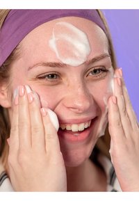 Schuimende gezichtsreiniger aangebracht op een glimlachend gezicht met een lichte huid en sproeten. Paarse hoofdband, handen met lichtroze nagels. Soepele textuur.