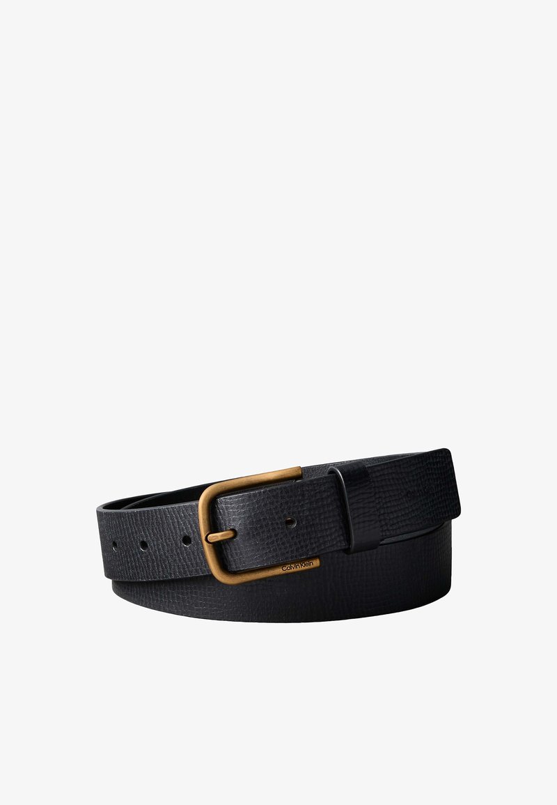 Cinturón de cuero negro con textura, hebilla de latón y logo de Calvin Klein grabado en el marco de la hebilla, enrollado sobre un fondo blanco.