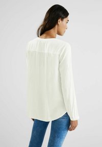 Blusa a maniche lunghe di un leggero color crema realizzata in tessuto morbido con orlo arrotondato, caratterizzata da un delicato dettaglio di cucitura sulla parte superiore della schiena.
