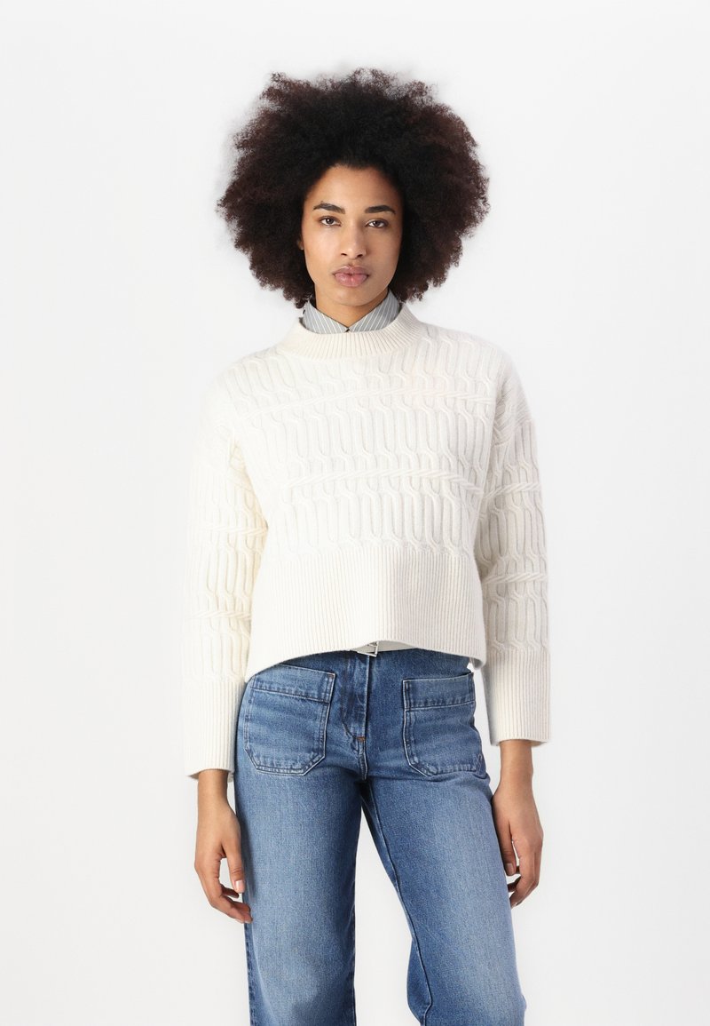 Pull en tricot couleur crème avec un motif texturé, doté d'un col montant et d'un design court, associé à un jean en denim bleu.