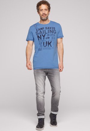 Uomo con capelli ricci e barba che indossa una t-shirt grafica blu, jeans grigi e scarpe da ginnastica nere, in piedi davanti a uno sfondo semplice.