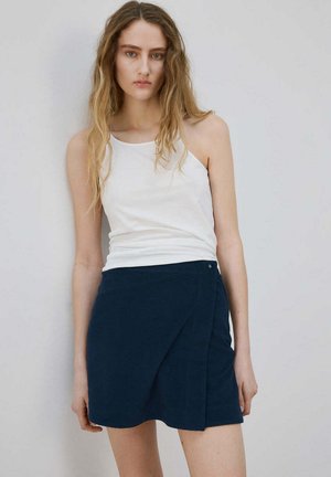 Joven con el cabello largo que lleva una camiseta blanca sin mangas y una falda azul oscuro, de pie frente a un fondo liso de color gris claro.