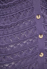 Cardigan tricotat de culoare violet, cu modele texturate ce includ motive de diamante și valuri. Șase butoane mari, de culoare deschisă, pe partea din față.