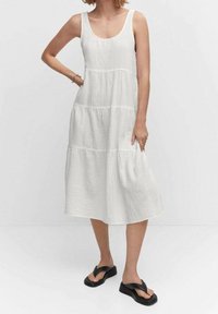 Abito estivo bianco, fino al ginocchio, con un design senza maniche, texture a strati e una finitura leggermente stropicciata. Abbinato a sandali flip-flop neri.