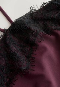 Violets atlas audums ar melnas ziedu mežģīņu apdari un plānām violetām lencītēm, kas izceļ tekstūru un smalkas detaļas.