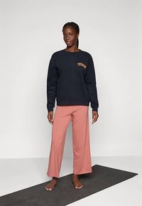 Felpa blu navy con testo arancione sul petto, abbinata a pantaloni larghi rosa chiaro, indossata a piedi nudi su un tappetino da yoga scuro.