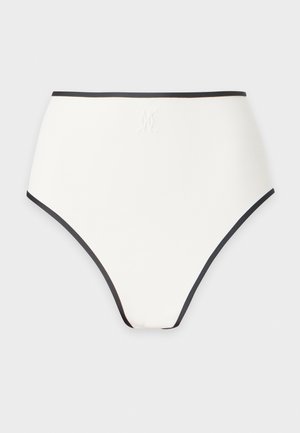 Femme debout en sous-vêtement blanc deux pièces avec bordures noires et sandales noires sur fond blanc uni.
