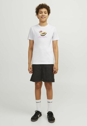 T-shirt bianca in cotone con stampa grafica colorata sul davanti, abbinata a pantaloni corti in denim neri e sneaker nere.