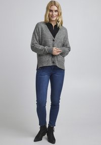 Grå cardigan med v-hals, knaplukning og frontlommer, parret med mørkeblå skinny jeans og sorte ankelstøvler. Glat tekstur.