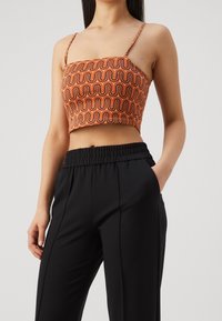 Kvinna i en orange mönstrad crop top med tunna axelband och höga svarta byxor med elastisk midja, handen i fickan.