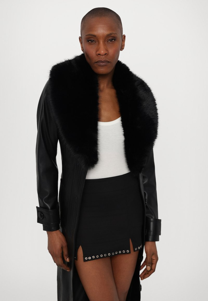Sort læderfrakke med en falsk pelskrave, lange ærmer og knappede manchetter, båret over en hvid tanktop og en sort miniskørt med grommet-detaljer.