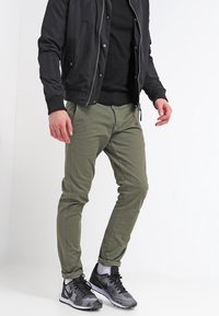 Olivově zelené slim-fit chinos vyrobené z bavlněné směsi, s zipovým zapínáním, bočními kapsami a minimalistickým designem. V kombinaci s černou bomber bundou a teniskami.