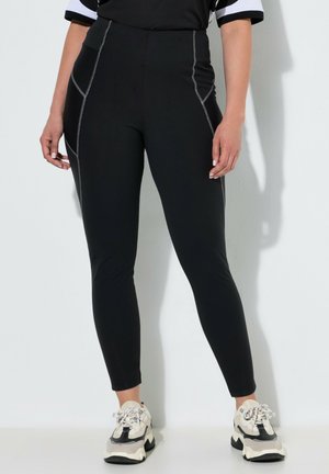 Kvinde iført sorte tætsiddende leggings med kontrastsyning og hvide chunky sneakers, stående mod en ensartet lys væg.
