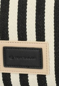 Sac fourre-tout rayé avec un motif chevron noir et crème, présentant un patch en cuir noir avec le logo et des bordures beiges. Matériau en toile texturée.