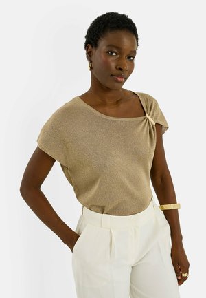 Mujer con cabello corto y rizado que lleva una blusa beige brillante, pantalones blancos de talle alto, pendientes de aro dorados y una pulsera y anillo dorados a juego.
