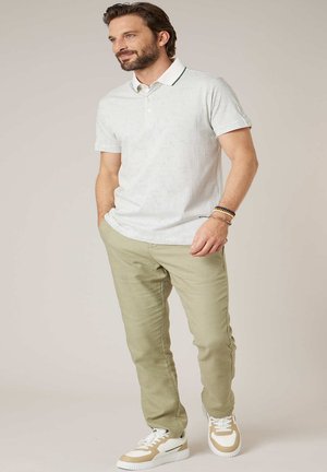 Homme portant un polo blanc à motifs, pantalon beige, baskets et bracelets, debout les mains dans les poches contre un fond neutre.