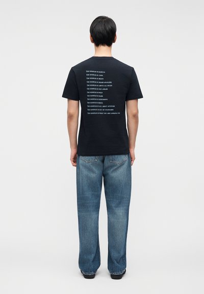 Μαύρο βαμβακερό t-shirt με επαναλαμβανόμενο λευκό κείμενο στην πλάτη, σε συν δυασμό με φαρδιά μπλε τζιν και μαύρα παπούτσια.