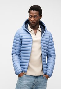 Άνδρας που φοράει ανοιχτό γαλάζιο γεμιστό μπουφάν με κουκούλα πάνω από μπεζ πουκάμισο με φερμουάρ, με τα χέρια στις τσέπες, κοιτάζοντας προς τα πλάγια σε απλό φόντο.