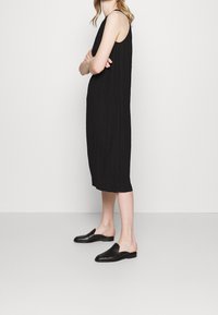 Musta, hihaton mekko polvipituudella ja teksturoidulla kankaalla. Yhdistetty tasapohjaisten mustien liukuvaatteiden kanssa.