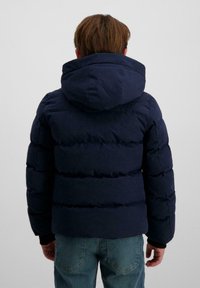 Kind van achter gezien, gekleed in een marineblauwe gewatteerde jas met capuchon en blauwe jeans, staand tegen een effen witte achtergrond.