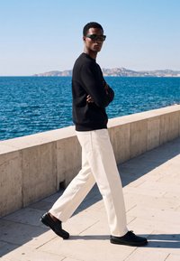 Homme portant un pull noir, un pantalon blanc et des chaussures noires marchant le long d'une promenade en bord de mer avec des montagnes en arrière-plan.