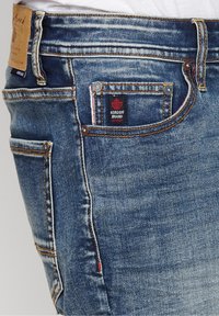 Denim jeans in dark blue with a classic fit. Features include orange stitching, a small red brand label on the front pocket, and rivets.