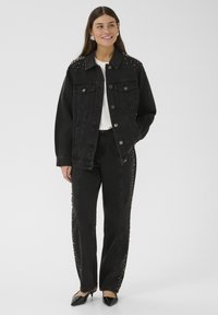 Žena koja nosi crnu jaknu od trapera sa čavlima i odgovarajuće pantalone, belu majicu i crne štikle, stoji s jednom rukom u džepu i smeška se.