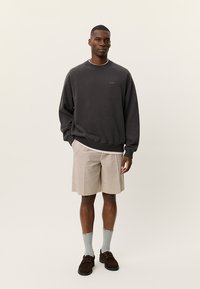 Ανδρας που στέκεται φορώντας σκουρόχρωμο γκρι φούτερ, μπεζ σορτς, ανοιχτό γκρι κάλτσες και σκούρα καφέ παπούτσια, μπροστά σε ένα απλό φωτεινό φόντο.