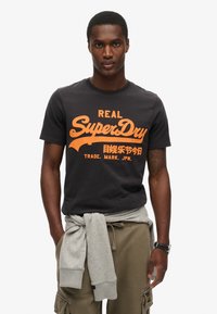 Camiseta de algodón negra con gráfico en naranja de texto "SuperDry", cuello redondo, mangas cortas, combinada con un sudadera gris atada a la cintura.
