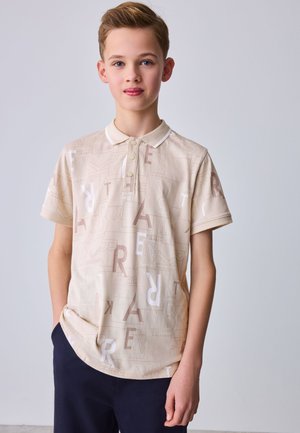 Jongen met lichtbruin haar, die een beige poloshirt draagt met verspreide letters en een donkere broek, staand tegen een effen lichte achtergrond.