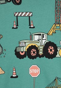 Zaļā audumā ir attēloti būvniecības transportlīdzekļi, tostarp dzeltenais traktors ar lieliem melniem riteņiem, satiksmes sarkanie konusi un apstāšanās zīme.