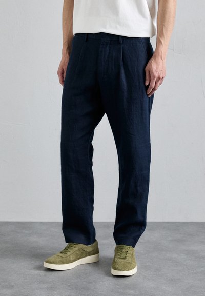 Marineblå linnedragt med forfald, parret med olivengrønne ruskinds sneakers med hvide såler. Glat tekstur og afslappet pasform.