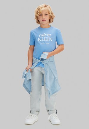 Lyshåret barn iført en blå Calvin Klein T-shirt, lyse denimjeans, hvide sneakers og en lys denimjakke bundet om taljen.