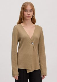 Beige wikk cardigan met een diepe V-hals, lange mouwen en een zilveren knoopsluiting. Zachte textuur en asymmetrische onderrand.