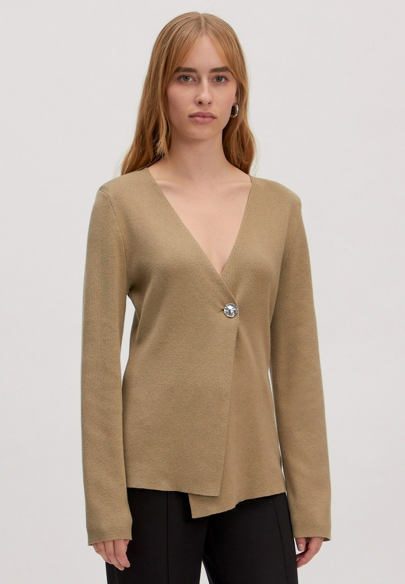 Beige wikk cardigan met een diepe V-hals, lange mouwen en een zilveren knoopsluiting. Zachte textuur en asymmetrische onderrand.