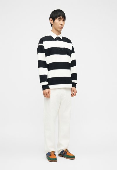 Άνδρας που στέκεται φορώντας polo πουκάμισο μακρυμάνικο με ασπρόμαυρες ρίγες, λευκό παντελόνι και πολύχρωμα παπούτσια σε αποχρώσεις του μπλε, πορτοκαλί και πράσινου.