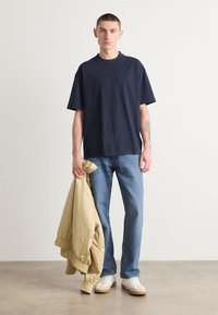 Námořnické modré bavlněné tričko s volným střihem, doplněné světle modrými džínami a béžovou bomber bundou přehozenou přes jeden rukáv. Bílé tenisky.