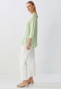 Blusa verde chiaro con maniche a tre quarti, morbida al tatto e con vestibilità rilassata. Abbinata a pantaloni bianchi a vita alta e sandali beige.