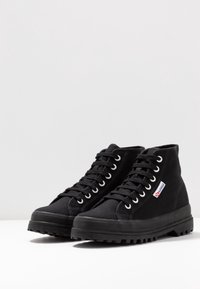 Black high-top canvas sneakers with metal eyelets, textured fabric, rubber sole, and prominent logo patch. Durable and versatile design.