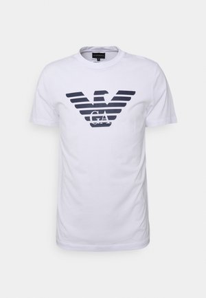 Hvit bomulls t-skjorte med en marineblå ørn-logo grafikk med striper og bokstavene "GA" i midten. Kort ermer og rund hals.